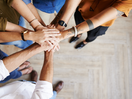 Diverse group of hands joined in a stack, showing teamwork. Wooden floor background, casual attire, tattoos, and watch visible. Positive mood.
