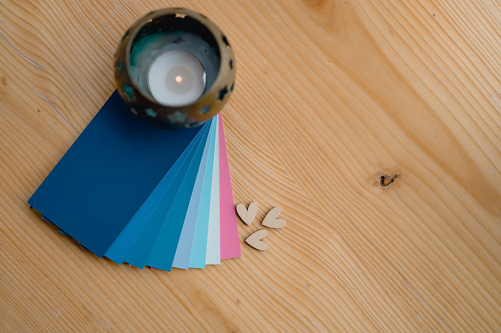 Candle in a holder on a wooden table beside fanned blue, pink, and white color swatches and small wooden heart shapes, creating a cozy, artistic vibe.