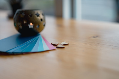 Front view of a candle in a holder on a wooden table beside fanned blue, pink, and white color swatches and small wooden heart shapes, creating a cozy, artistic vibe.