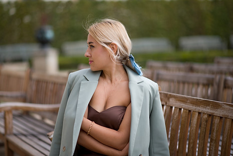 Blonde woman wearing brown dress and blue coat, sitting on a bench in Wallenstein Garden in Prague at Martina Bromkova Photography