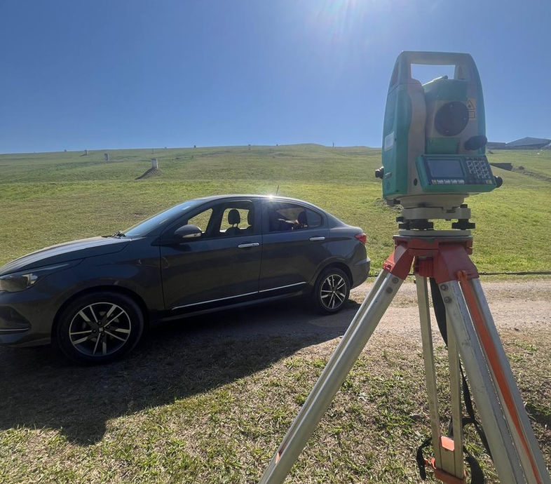 Estação Total topográfica montada em tripé em um campo aberto ensolarado, com o sol ao fundo e um carro da equipe próximo