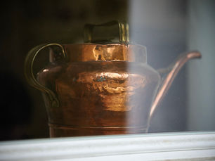 A hammered copper vessel with a handle, a lid and a long spout, photographed through window glass with a white sill below