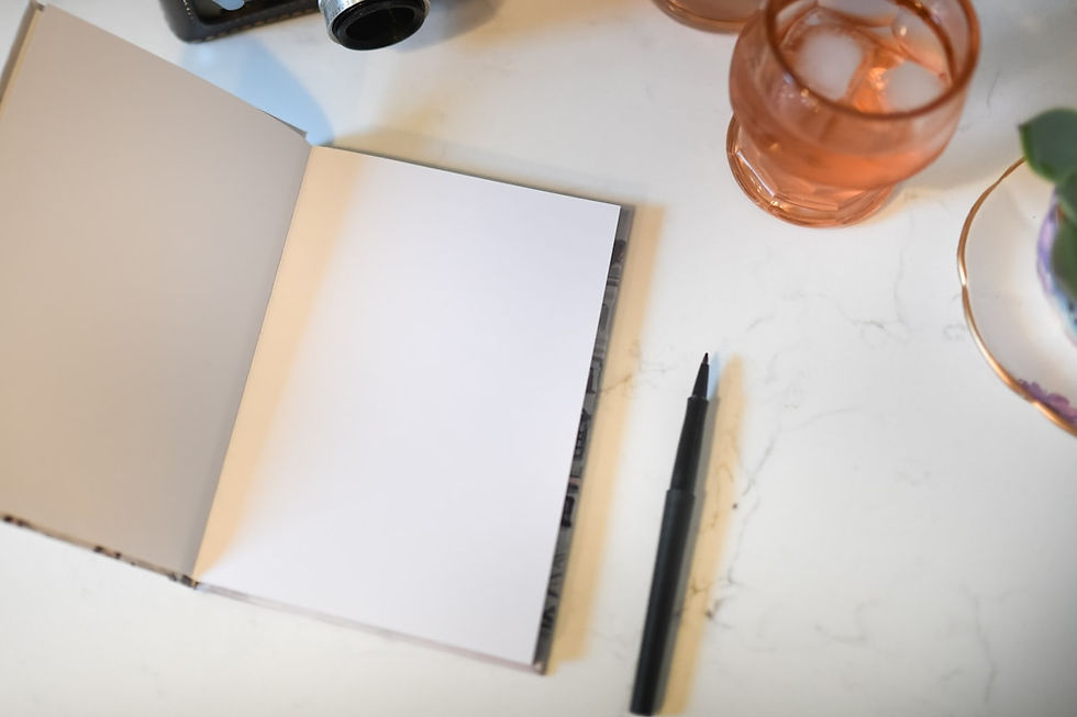 An open blank notebook with a black pen resting beside it on a pale marble surface, with a rose tinted drinking glass at the top edge of the frame