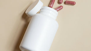 A plain white unlabeled supplement bottle tipped on its side on a beige background, cap removed, with several dark red capsules spilled out beside it