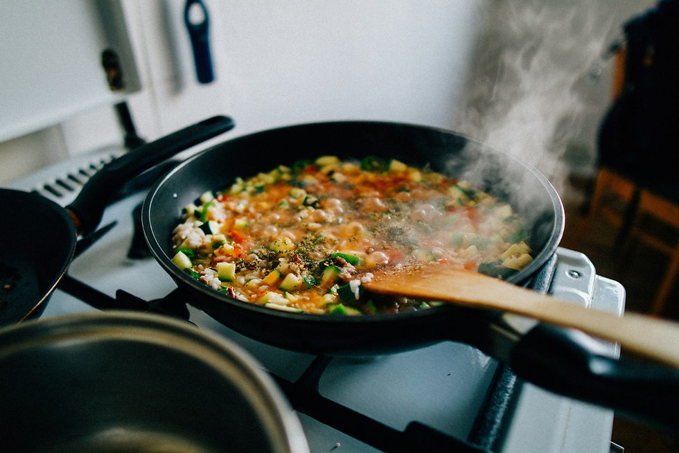 A dark pan of chopped vegetables and rice simmering on a stovetop with visible steam rising and a wooden spoon resting in the pan