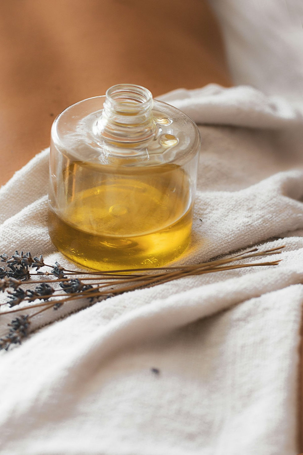 A small open glass bottle of golden oil on a pale woven cloth, with dried flower sprigs and thin dry stems lying beside it