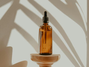 An amber glass dropper bottle of golden oil standing on a small turned wooden stand against a cream wall, with palm frond shadows falling across it