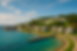 A bright, realistic coastal scene inspired by Mackinac Island, showing pastel-colored houses with porches along a curved shoreline, a white church with a steeple, a small dock extending into clear turquoise Great Lakes water, and a historic fort on a hill surrounded by lush green trees under a clear blue sky.
