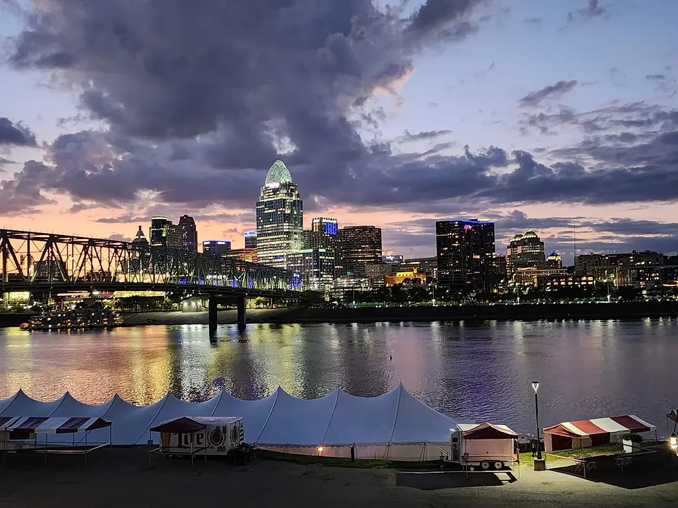 Cincinnati skyline at dusk viewed across the Ohio River, with illuminated downtown buildings, a bridge spanning the river, and reflections of city lights on the water under dramatic evening clouds.