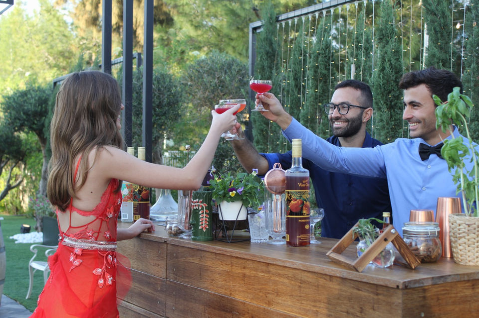 שלושה אנשים שמחים מרימים כוסית קוקטיילים בבר עץ חיצוני.