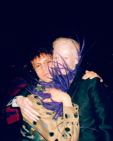 Flash Photograph of Julio Torres and Tilda Swinton photographed by Daniel Roa. Tilda is wearing a large purple feather neckpiece and Julio is wearing a golden polka dot top. 