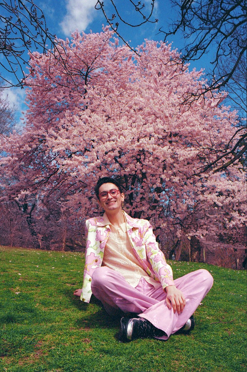 Photographer Daniel Roa in colorful clothes sits on grass, smiling in front of a blooming cherry blossom tree under a blue sky. The scene is vibrant and joyful. photographed on 35mm film.