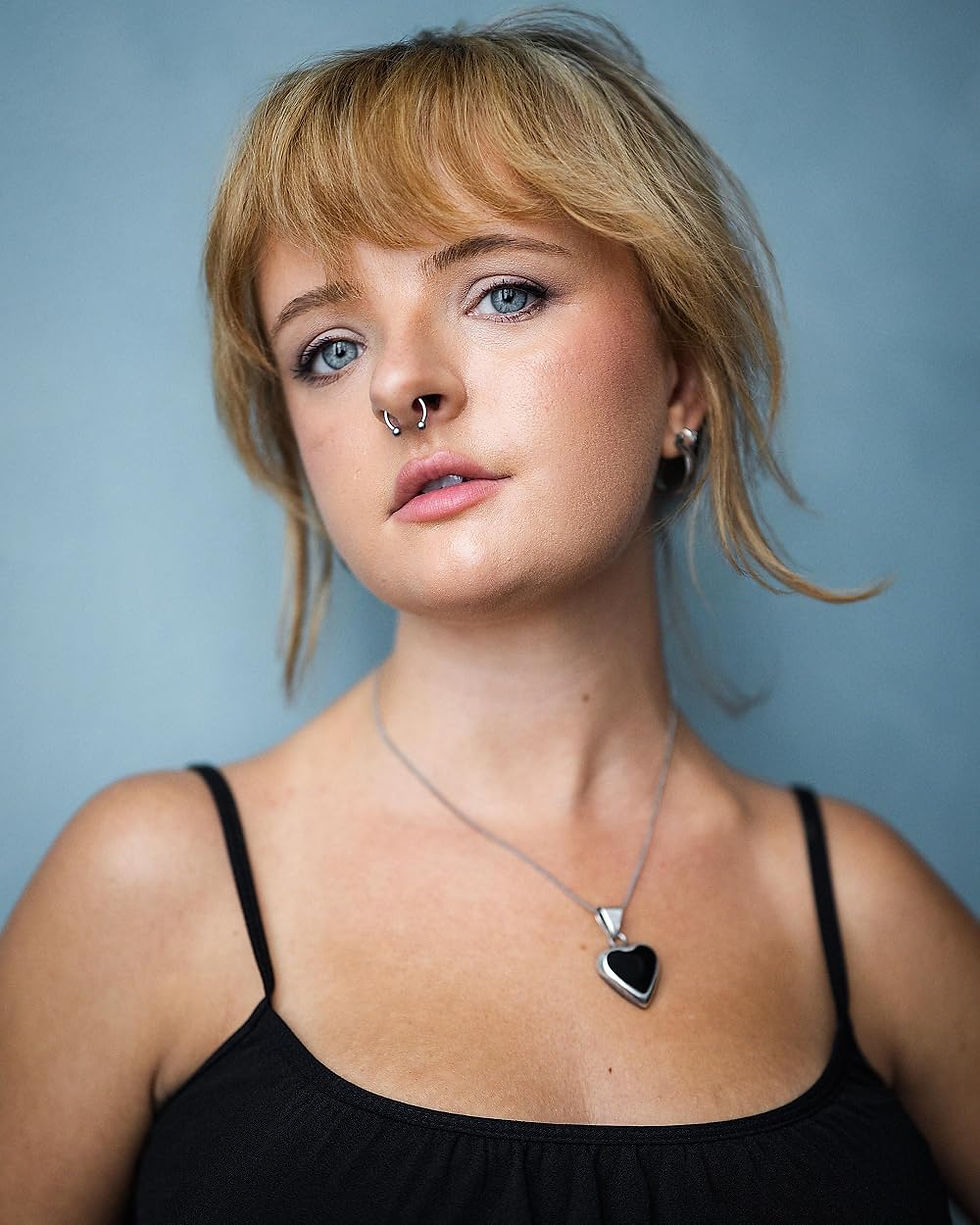 Actress Milly Shapiro in a professional photo with a grayish blue background, wearing a black tank top, silver septum ring, and a black, heart-shaped necklace.