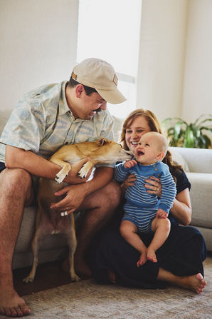 A laughing family of three, a baby boy and his parents, and a dog on the sofa