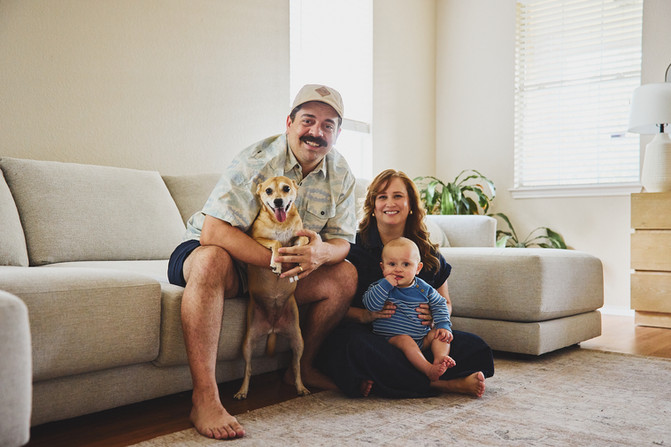 A laughing family of three, a baby boy and his parents, and a dog on the sofa