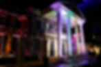 A mansion at night is illuminated with colorful lights—purple, green, and pink. Two people walk nearby, creating a festive atmosphere.