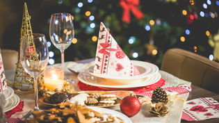 Festive table set with Christmas-themed napkins, a lit candle, cookies, and a red ornament. Blurred lights of a decorated tree in the background.