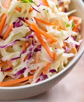 A white bowl containing coleslaw made from grated carrots, red cabbage, white cabbage, and a vinegar dressing. It sits on a wooden cutting board.