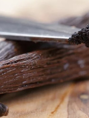 A close-up of a vanilla bean with a knife tip that has vanilla seeds in the tip. It sits on a wooden counter.