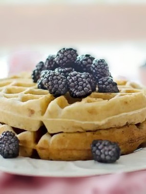 A close-up of freshly baked waffles with fresh blackberries on top.  They are on a white plate that sits on a pink dishtowel.