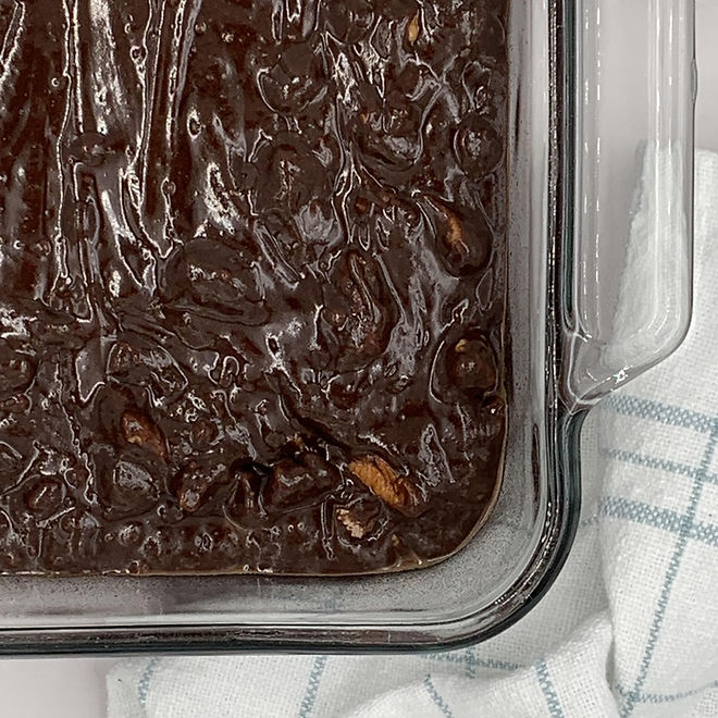 A close-up view of chocolate brownie batter in a square clear glass baking dish. The batter has a smooth, glossy surface with visible pieces of pecan nuts. The dish sits on a plaid blue and white dish towel.