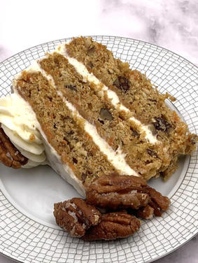 A 3-layered slice of carrot cake with cream cheese frosting garnished with candied pecans. It sits on a silver and white plate on a marble counter. 
