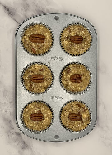 A silver muffin pan holds the batter of 6 pecan pie muffins. The muffins have a pecan half on top and the pan sits on a white marble counter.
