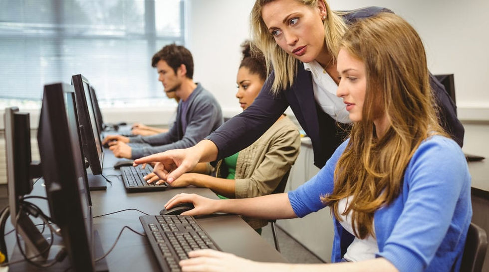 A teacher assists a student in a computer lab, providing guidance as others focus on their work.