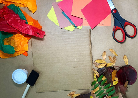 Image of a workspace for the fall collage free craft, with a square of cardboard, tissues paper, glue, a brush, scissors, colored paper, fall leaves all laid out