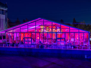 Nighttime outdoor event production with glass tent, colorful lighting, and lounge design