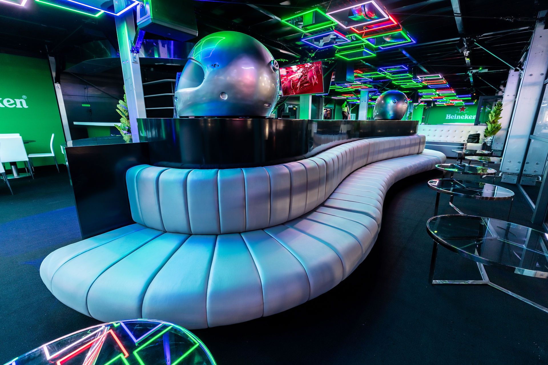 Heineken-branded lounge with neon ceiling lights, curved metallic sofa, and F1 helmet decor