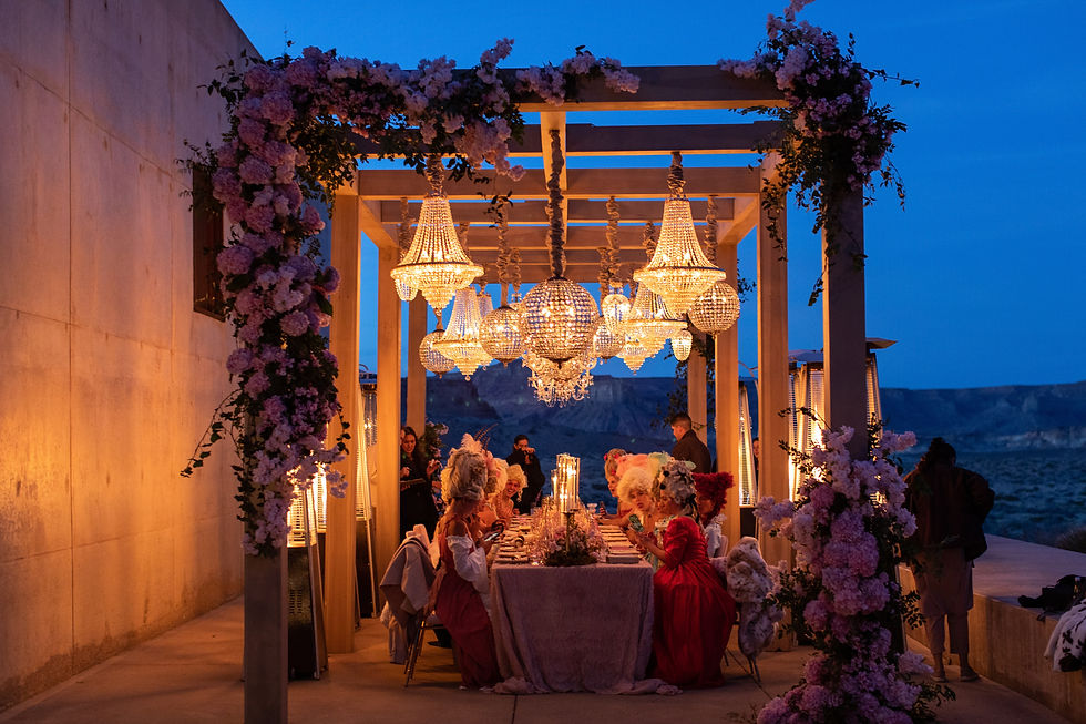 Evening tablescape under a pergola adorned with chandeliers and cascading pink florals, with guests in elaborate historical costumes.
