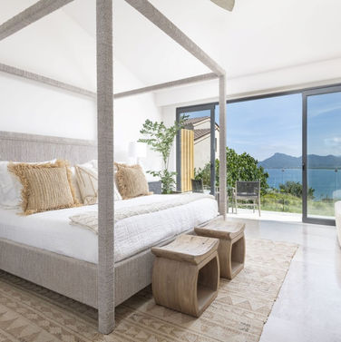 Luxury Vacation Home Serene Master Bedroom with Natural Woven Textures, Wood Furniture Accents, White Walls and a View of the Ocean off the Balcony
