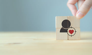 The image shows a simple, symbolic scene built around four small wooden blocks arranged into a square. On the front of the blocks is a minimal icon of a person (a black silhouette), with a red heart positioned over the chest area. A circular target or crosshair overlays the heart, emphasizing it as a focal point. A hand is gently placing or adjusting the top-right block, suggesting intentional positioning or selection.