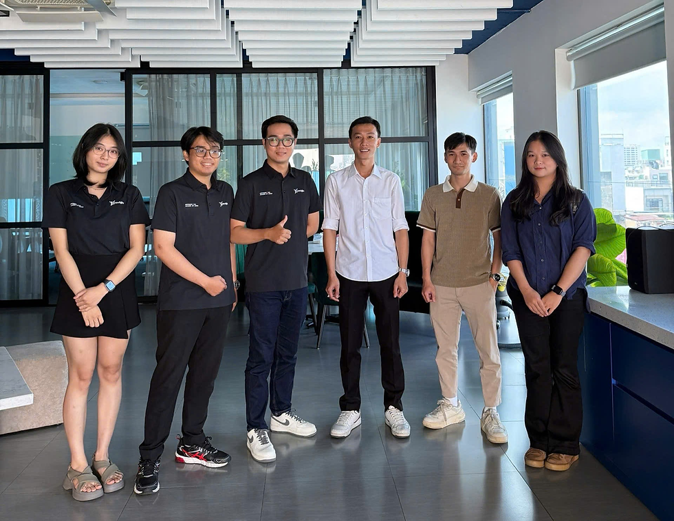 Six people standing indoors, smiling, in a modern office. Three wear matching black shirts, others in casual attire. Bright, professional setting.