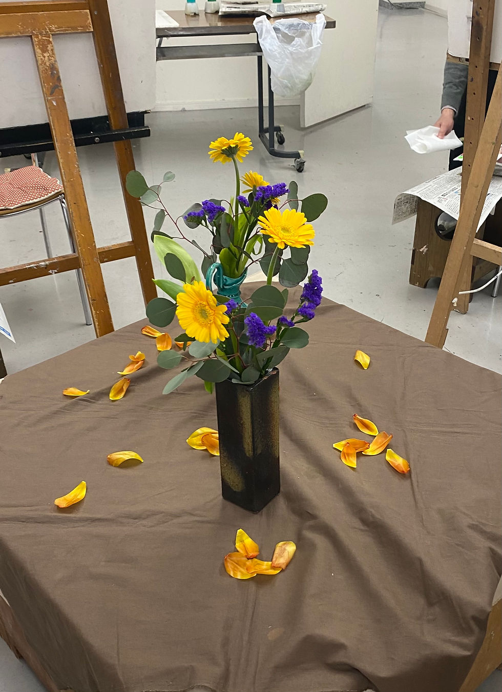 茶色い布をかけた台の上に、黒い花器の黄色いガーベラと紫の花の生け花。周囲に花びらが散り、制作中のアトリエの雰囲気】【。