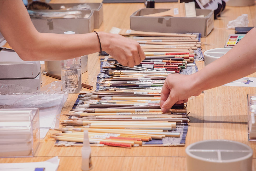 木製テーブルの上で、2人の手が並べた絵筆を選別している。箱や容器が周囲にあり、作業中の落ち着いた雰囲気。