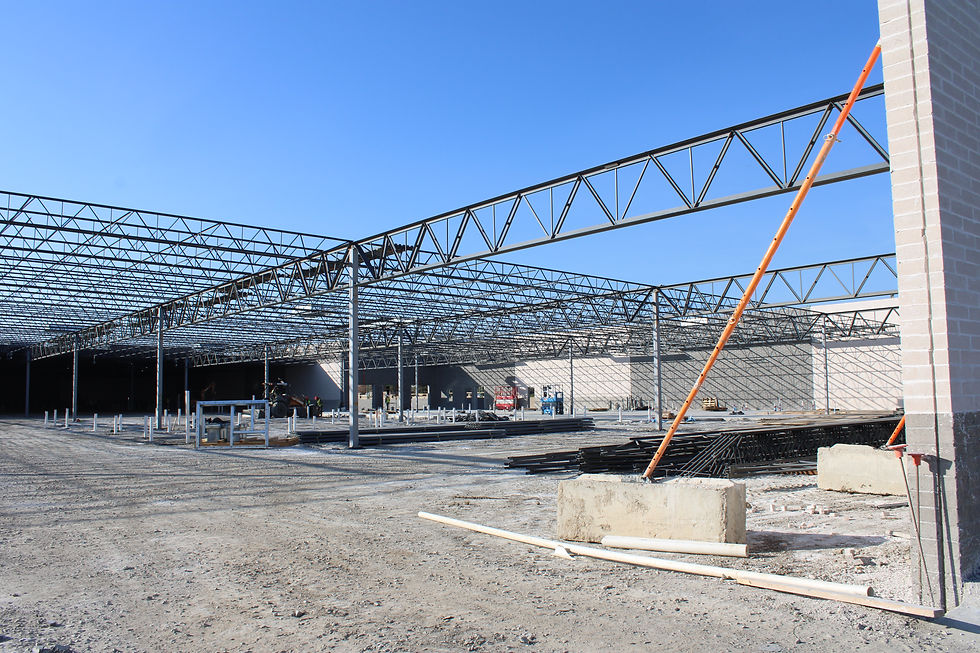 Steel roof trusses rise over a construction site under a clear blue sky, with an orange brace, concrete blocks, and scattered materials.