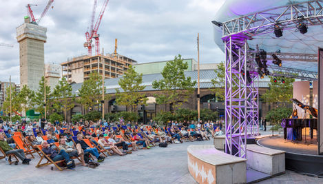 An audience in Lewis Cubitt Square, King's Cross, sat watching a pianist perform as part of Cubitt Sessions, on a temporary stage to the right of the image.