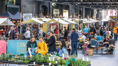 A busy scene of people in the West Handyside Canopy at King's Cross, in front of Canopy Market stalls. People stand in the foreground browsing plants, with other people browsing and buying food and drinks from stalls, and sat at tables in the background.