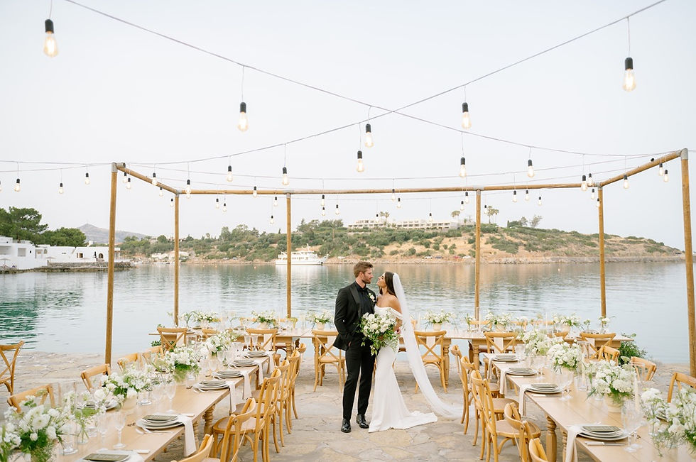 Sea side wedding reception in Crete, wooden tables with floral arrangements, string lights above, beautiful wedding couple