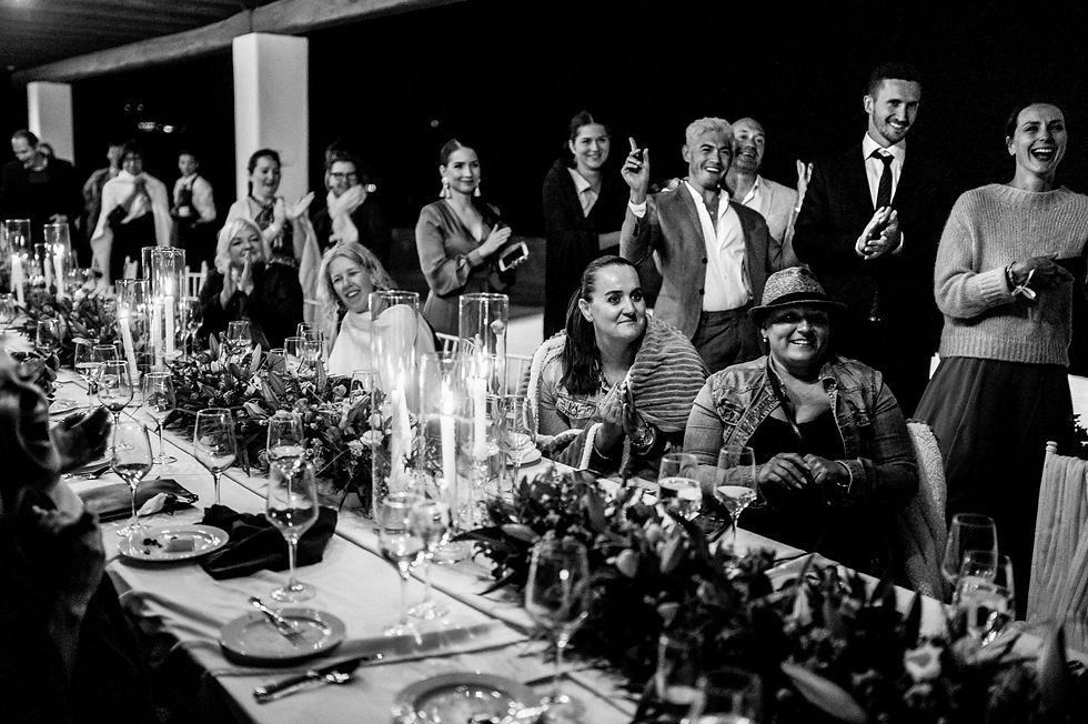 Guests seated and standing around a candlelit table at an elegant gathering, clapping and smiling in a joyful, festive atmosphere. First dance reaction.