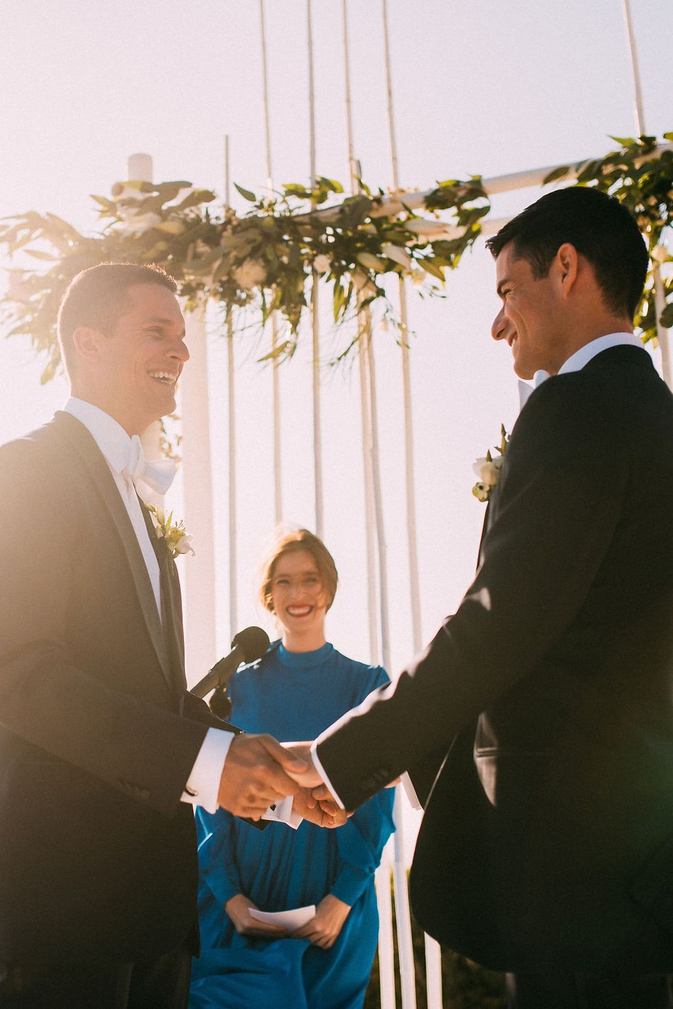 Two men in suits hold hands, smiling, during an outdoor wedding ceremony. A woman in blue officiates. Greenery decorates the background. Aisle. Arch. Same-sex marriage. Mykonos. 