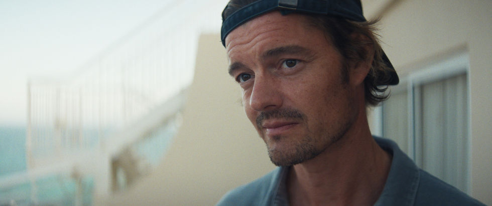 Man wearing a backward cap gazes pensively off-camera. Blurred outdoor background with stairs and ocean, conveying a calm, introspective mood.