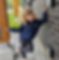 Stella Pasqua is smiling while wearing a Meaningful Access hoody. Her hair is blond, and she is standing, crossing one leg in front of the other. She is outside near an automatic door button, leaning against a brick wall.