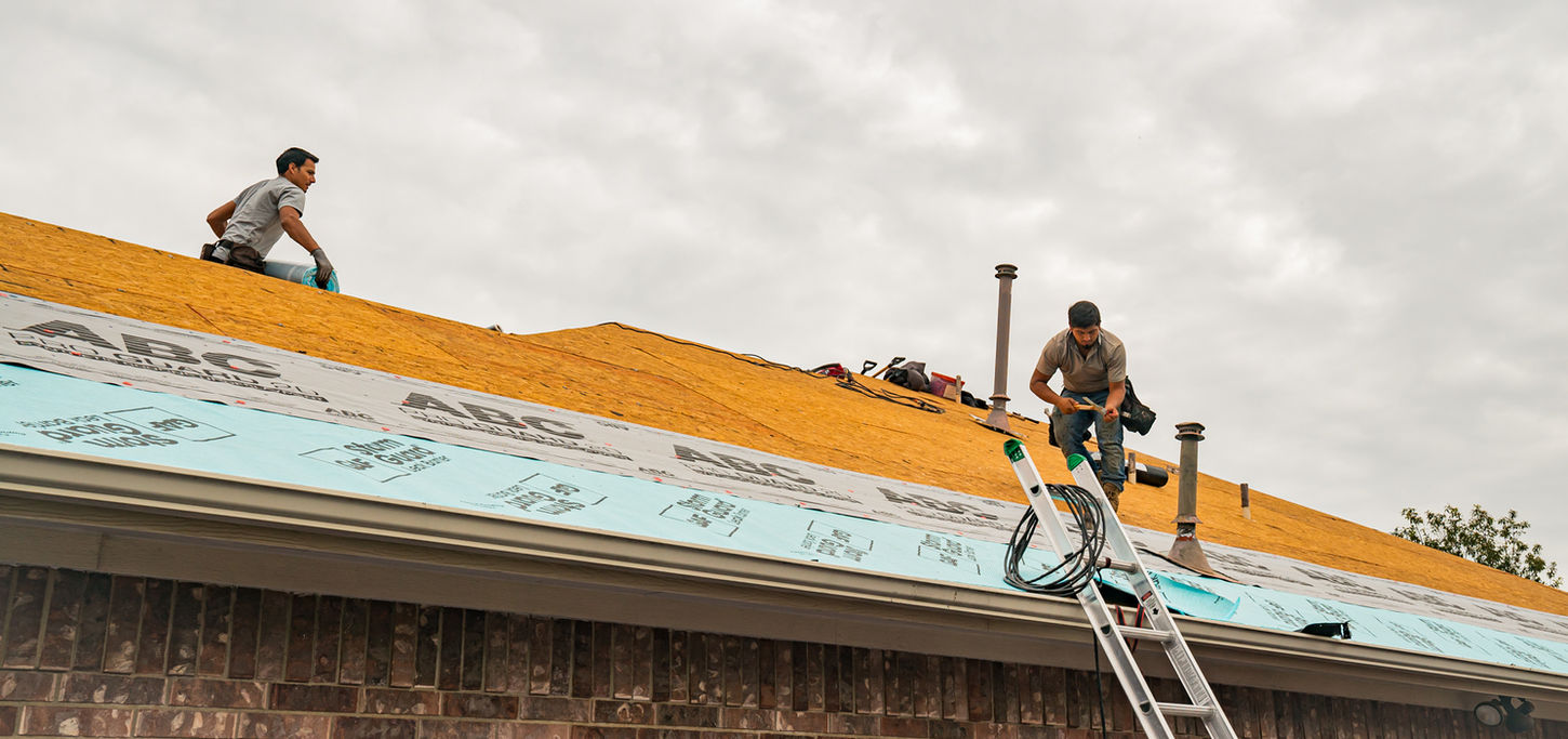 Two workers on a roof, one installing synthetic underlayment while the other holds a roll of ice and water shield, preparing for the next step.