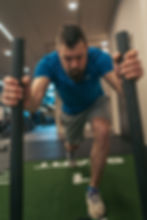 Mann beim Push-Sled-Training im Fitnessstudio – Er trägt ein blaues Funktionsshirt und graue Shorts und schiebt mit konzentriertem Blick einen Gewichtsschlitten über eine grüne Kunstrasenbahn mit der Aufschrift ‚START‘. Die Szene vermittelt Stärke, Ausdauer und intensives Fitnesstraining.