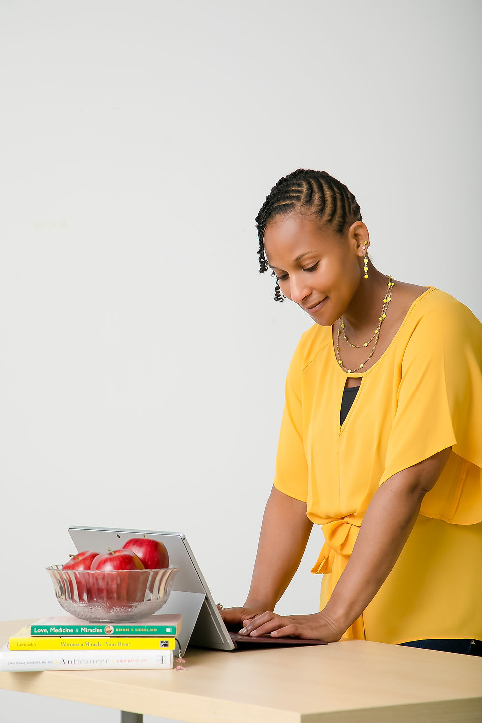 A woman in a yellow top types on a laptop at a table with books and apples. Bright, simple background; focused mood.