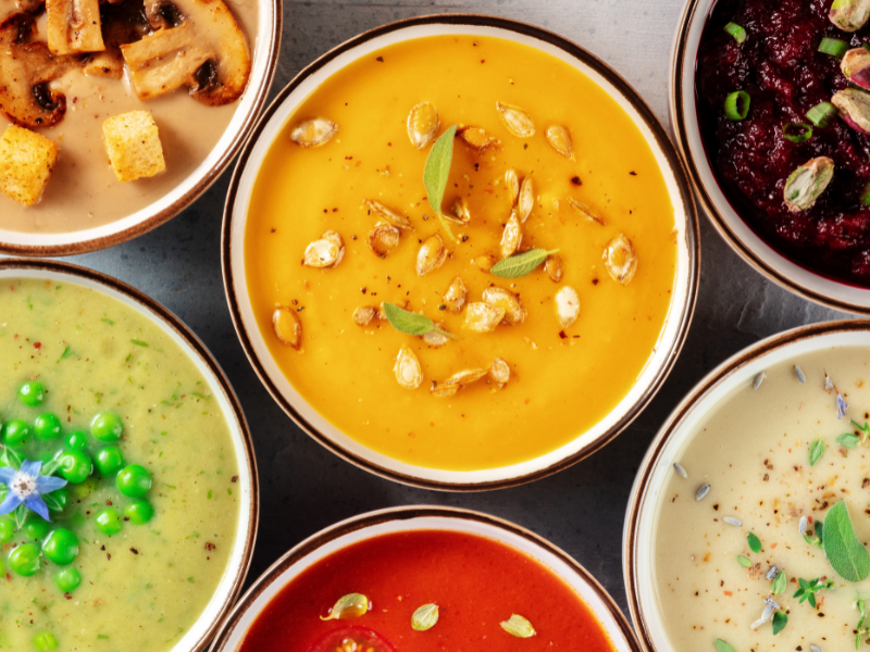 Colorful bowls of soup in various flavors, topped with garnishes like seeds, mushrooms, peas, and herbs, arranged on a gray surface.