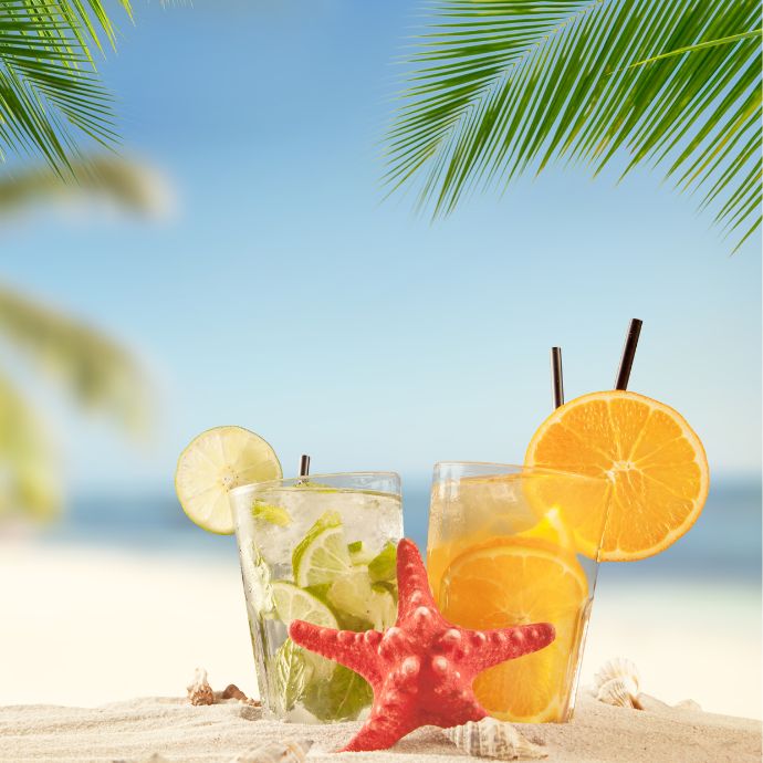 Two tropical drinks with lime and orange slices sit on sandy beach, accompanied by a red starfish and palm leaves overhead. Two tropical drinks with lime and orange slices sit on sandy beach, accompanied by a red starfish and palm leaves overhead.